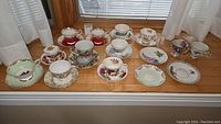 Full display of 13 assorted vintage tea cups and saucers laid on wood surface under window