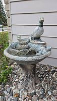 Full view of the heavy cement outdoor electric water fountain showing the duck figure on top and the textured column base among garden stones.