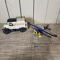 Both vintage Coca Cola vehicles on wood surface in front of white wood paneled wall. Truck is white cast iron with Coca Cola logo, plane is blue diecast with yellow propeller and landing gear.