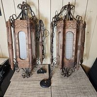 Front view of both vintage hanging lights showing carved wooden panels with floral designs, vertical carved spindles, glass panes with grid, iron scrollwork, metal chains and bases.