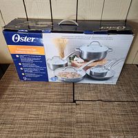 Outer box showing Oster brand and visual of the cookware set including pots and pans with lids.