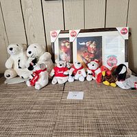 Overview of all Coca-Cola plush bears, small plush birds and penguin, along with two framed Coca-Cola plaques showcasing vintage art.