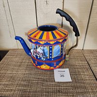 Side view of colorful carousel horse-themed tea kettle showing blue spout, black handle, and decorative horses.