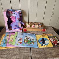 Photo showing vintage purple Barbie Dragon plush toy in original packaging, several vintage Little Golden Books including Donald Duck titles, wooden alphabet blocks on red wagon, and additional children's books.