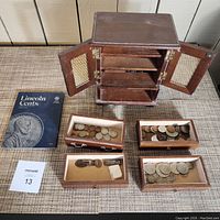 Full view of the wooden musical jewelry box with doors open revealing three shelves and four small drawer trays with coins on surface next to it along with Lincoln cents folder.