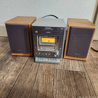 Photo showing the Panasonic stereo system main unit flanked by two wooden speakers on a wooden floor, unit powered on with display lit