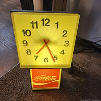 Vintage Coca Cola advertising wall clock with square yellow clock face, black numerals, red hands, and illuminated 'Enjoy Coca Cola' panel beneath. Clock's plastic cover cracked near logo.