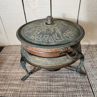Top view of the vintage hammered copper pot with lid showing camel and Middle Eastern designs on lid and metal footed stand underneath.