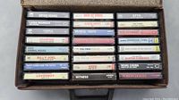 Cassette tapes arranged in a carrying case, showing artist and album titles on tape labels including Bryan Adams, Styx, Rod Stewart, Loverboy, Billy Joel, and others.