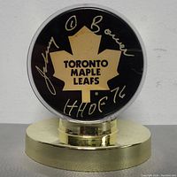 Front view of signed Toronto Maple Leafs hockey puck showing Johnny Bower autograph and HOF 76 inscription in gold marker, inside clear plastic case on gold stand.