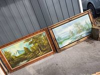 Two landscape paintings leaning against a gray wall outdoors showing mountains, trees, and water with ornate gold-toned frames.