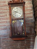 Full view of wooden wall clock with decorative carved top, turned columns, glass door with gold accents, and clock face with Roman numerals.