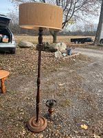 Tall wooden floor lamp with turned stand and beige fabric shade, shown standing outside on gravel and leaves.