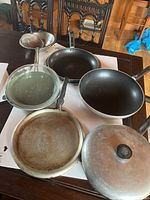 Six miscellaneous pans on a table, including non-stick frying pans, a glass pan, and metal pans showing use wear