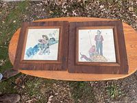 Two wooden framed Norman Rockwell prints displayed on a wooden tabletop outdoors showing autumn leaves on the ground.
