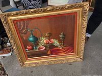Framed still life painting showing fruit and decorative vessels on a table, signed by artist.
