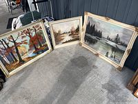 Three framed landscape paintings resting on the ground showing different seasonal scenes, including autumn trees and rivers.