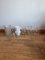 Seven vases arranged in a row including six clear glass and one white opaque textured vase.