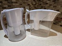 Two Brita water filter pitchers shown side by side on a kitchen counter, clear containers with white tops and handles, internal filter compartments visible.