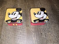 Two small square metal watch boxes with colorful Mickey Mouse artwork on lids shown closed on a wood surface.