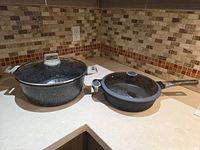 Both ceramic casserole pot and frying pan with lids, displayed on countertop against tiled backsplash.