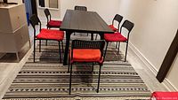 Dining table set with black IKEA dining table and six black perforated metal chairs with red cushions on a striped black and beige rug.