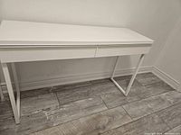 Front angled view showing off-white wooden desktop and white metal legs of computer desk on gray wood floor.