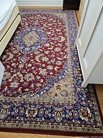 Full view of the Persian-style rug under a bed and near a white wardrobe. Highlights the central medallion and floral pattern in red, blue, and beige tones.