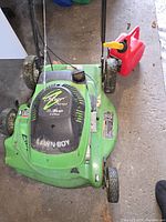 Photo showing top-down view of green Lawn Boy mower with silver series 4.5 HP 4-cycle engine label and black wheels. Red gasoline dispenser with yellow spout next to it.