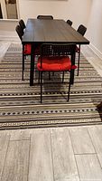 Photo of a black dining table with six black chairs with red cushions, set over the Persian-style Klim rug. The rug features multiple horizontal stripes with geometric patterns, in muted colors including beige, black, gray, and white.