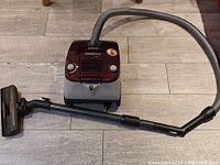 Front view of the vacuum cleaner showing the body, hose, extendable black rigid tube and rectangular floor brush attachment on tile floor.