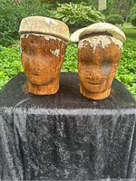Front view of two wooden hat blocks with sculpted human faces and cracked paint on domes