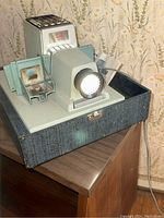 Photo showing the front and top view of the Argus 500 slide projector resting inside its original carrying case with light on.