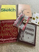 Photo of calligraphy books, two sketchbooks, calligraphy pens in plastic sleeve, and nib box.