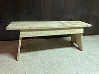 Front angle view of painted wooden farmhouse bench showing metal reinforcement under legs on linoleum floor and dark wood wall background.