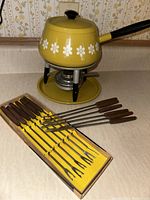 Photo showing yellow fondue pot with white flower decoration on stand and burner, with 12 brown-handled fondue forks in box