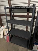 Front view of black resin four-tier shelving unit with textured grid pattern shelves, placed against metal wall and surrounded by household items.