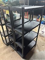 Black resin shelf with four shelves assembled with four tubular posts, photographed indoors on a concrete floor amid other items.