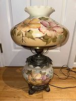 Full view of floral hurricane lamp showing glass base and shade with floral painting and metal base on wooden floor.