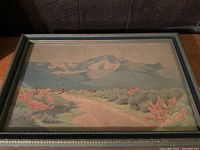 Framed watercolor landscape painting showing a mountain range with pink shrubbery and a dirt road leading into the distance.