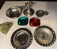 Photo showing all items in the lot on a speckled countertop: Gorham sterling weighted candlestick, two colored dishes (green and red), two wavy edged stainless steel dishes (one with glass insert), carved stone dish, iron ashtray.