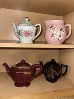 Top image showing two shelves inside a cabinet, each holding pottery. Top shelf contains a white floral teapot and a pink floral pottery pitcher. Bottom shelf has two dark pottery teapots, one brown and one black decorated with floral patterns.