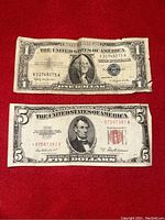 Two US currency notes placed on a red surface showing the one dollar silver certificate on top and the five dollar red seal note below.