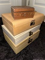 Stack of four jewelry boxes: small wooden carved box with a key on top, medium beige engraved box, two white rectangular boxes below.