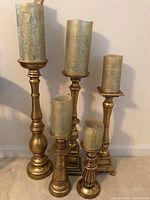 Five gold-tone resin candlesticks with faux candles arranged together against a wall on a carpet floor.