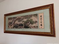 Framed rectangular embroidered artwork showing Chinese river scene with boats, bridge, trees, and people, with calligraphy on the right side. Mounted with a light green mat in a brown wooden frame.