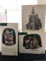 Photo of the three Department 56 ceramic buildings boxes: Staghorn Lodge, Teaman & Crupp China Shop, and Old Michael Church, showing front and side views of the packaging with detailed building photos and descriptions.
