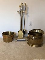 Full view of brass fireplace tools with white marble handles on a marble base, flanked by two brass ash buckets