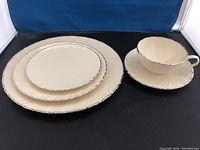 Three plates of different sizes stacked (dinner, salad, cake) along with a matching cup and saucer featuring ivory color with a scalloped edge and thin blue trim, showing the pattern detail.