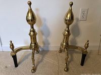Pair of Virginia Metalcrafters Williamsburg brass fireplace andirons placed on carpeted floor, showing front and side views.